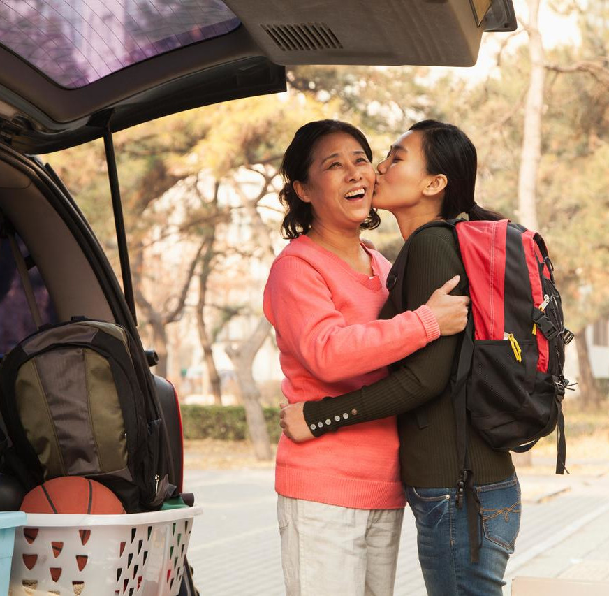 A mom and daughter embrace at the back of a car packed with belongings. 